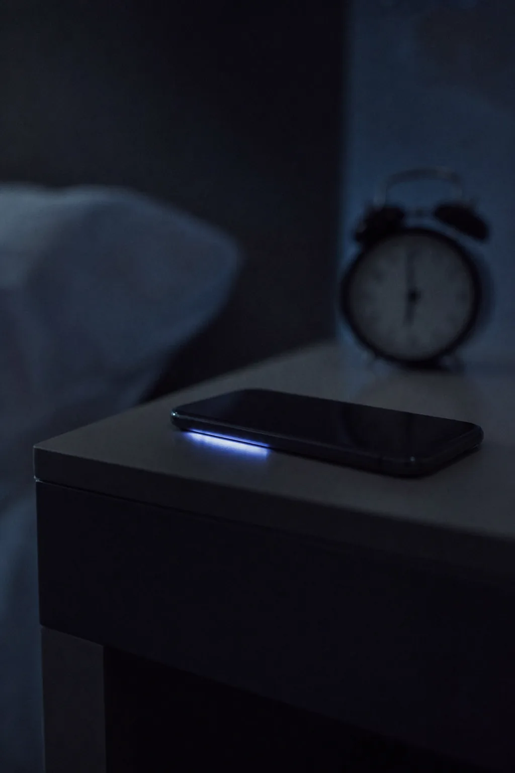 A phone face down on a bedside table in a dark bedroom, its edge glowing from a call.